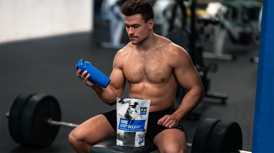 En mand sidder i et fitnesslokale med en pose Core Whey Delight og er ved at lave en protein shake. En mand sidder i et fitnesslokale med en pose Core Whey Delight og er ved at lave en protein shake.