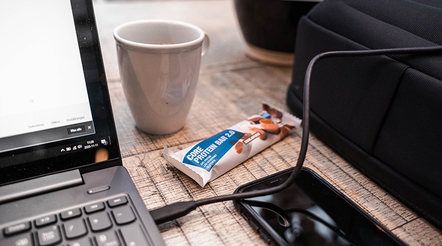 En Core Protein Bar 2.0 ligger p et skrivebord ved siden af en computer, kop kaffe og telefon.